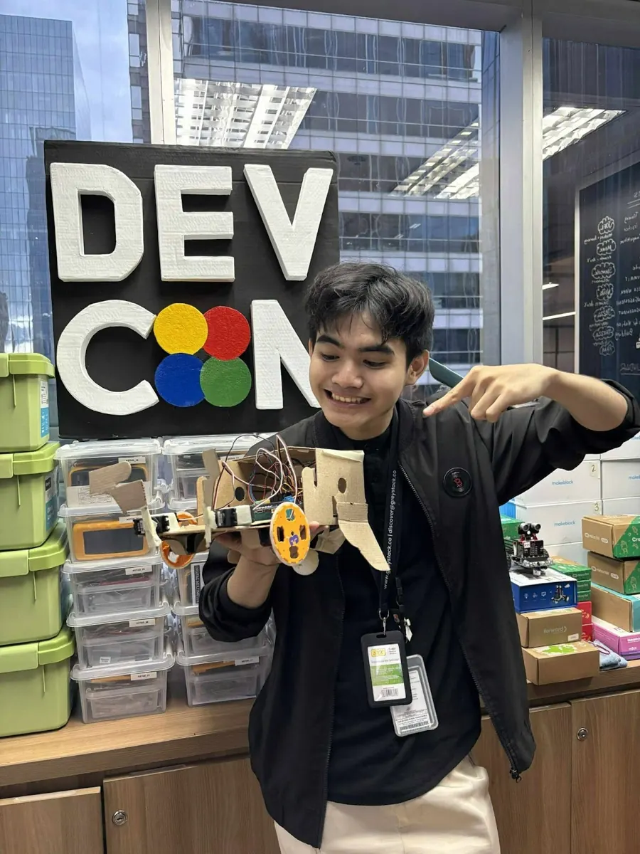 An intern proudly showing a cardboard robot rover he built, in front of the DEVCON sign
